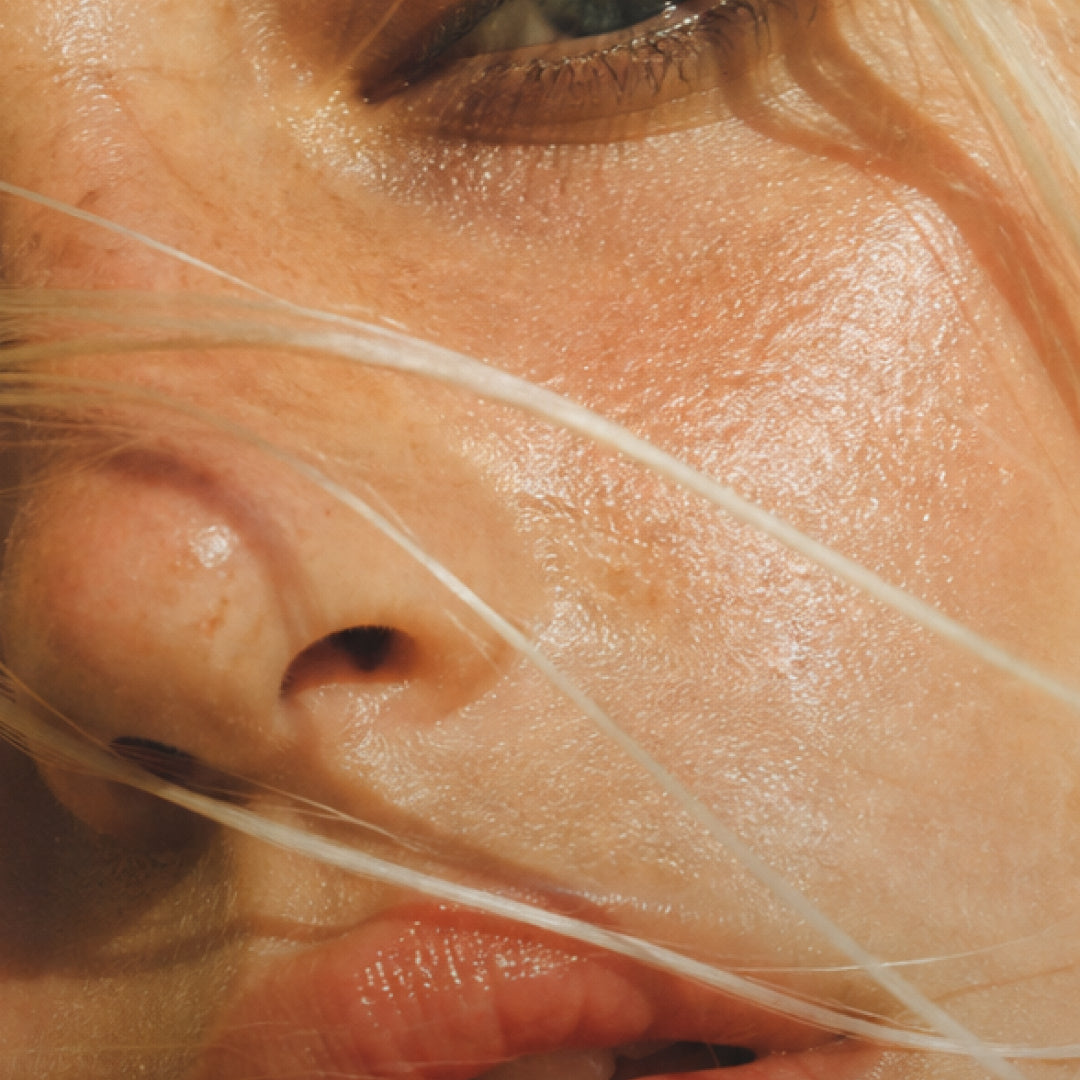 Extreme close-up of a person's face showing textured skin, a nose, lips, and a visible eye, with strands of light hair crossing the frame.